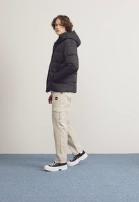 Padded black jacket with hood, beżowe spodnie cargo oraz czarne sneakersy z białymi podeszwami. Model stoi na niebieskim dywaniku na jasnym tle.