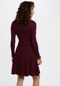 Vestido de canalé burdeos con mangas largas, cuerpo ajustado y falda plisada. Presenta una cintura entallada para una silueta definida.