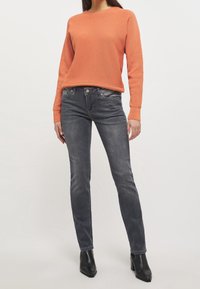 Suéter texturizado naranja con cuello redondo, combinado con jeans ajustados de un gris oscuro y botines negros, que exhiben líneas limpias y una silueta delgada.
