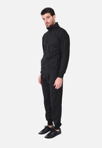 Giacca nera con zip e colletto alto, con tasche laterali, abbinata a pantaloni jogger coordinati. Completata con sneakers nere a suola bianca.