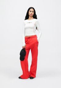 Bijela majica s dugim rukavima s crvenim naglascima i logotipom, uparena s crvenim trenirkama koje imaju elastični pojas i džepove. Crna kapa drži se u ruci.