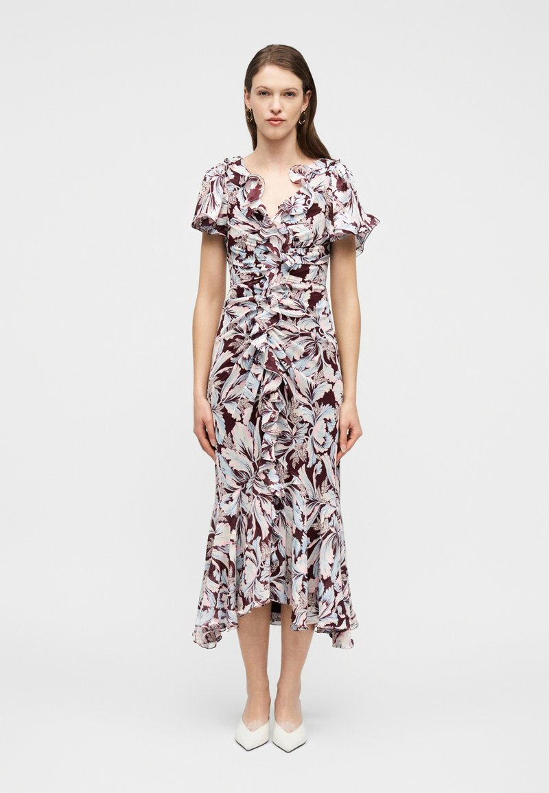 Γυναίκα που στέκεται, φορώντας ένα μίντι φόρεμα  μπορντό και ανοιχτό μπλε με λουλουδάτο σχέδιο, διακοσμημένο με βολάν, και λευκά μυτερά τακούνια, μπροστά σε απλό φόντο.