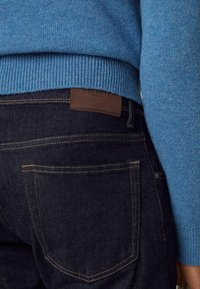 Blaues Strickpullover mit geripptem Saum, kombiniert mit einer dunklen Jeans aus Denim mit gelben Nähten und einem braunen Lederpatch mit der Aufschrift "Hackett London."