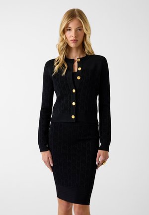 Blond kvinde iført en sort tætsiddende nederdel og matchende sort cardigan med guldknapper, stående mod en hvid baggrund.