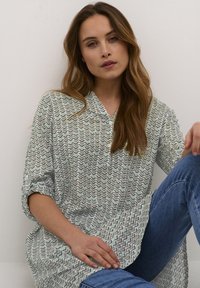 Tunique verte claire avec un motif en zigzag, patte de boutonnage, et manches retroussées, associée à un jean bleu. Tissu doux et coupe décontractée.
