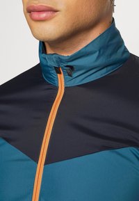 Herrejakke med høj krave, i marineblå med teal-detaljer, en orange lynlås og et glat, let materiale.