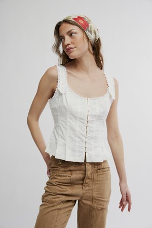 Giovane donna con indosso una camicetta bianca senza maniche con dettagli a bottoni e pantaloni marroni, che guarda di lato con un foulard floreale in testa.