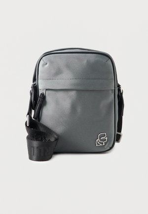 Kompaktná sivá kabelka cez rameno s predným vreckom na zips, čiernym nastaviteľným remienkom a malým strieborným logom v pravom dolnom rohu.