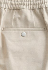 Tissu beige avec une ceinture élastique, une poche passepoilée horizontale et un unique bouton rond argenté centré sous la poche.
