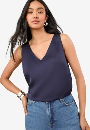 Mørkeblå satin top med V-hals og uden ærmer, parret med højtaljede denim shorts. En sølv armring på håndleddet tilføjer detaljer.