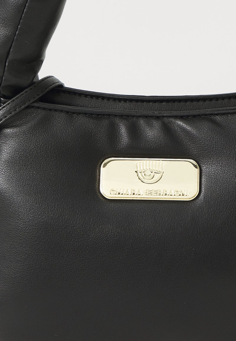 Sac à main en cuir noir avec une texture lisse, une anse courbée et une plaque logo rectangulaire en or affichant "Chiara Ferragni."