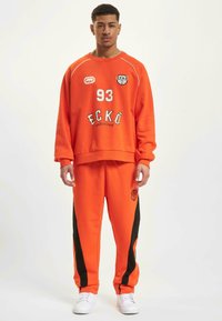 Orange sweatshirt and matching joggers with black side stripes, featuring front graphics and the number "93." Made of soft, textured fabric.