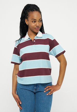 Tânără cu părul împletit, purtând un tricou polo cu mâneci scurte, dungat în albastru și brun-maroniu, și blugi albaștri, stând cu mâna pe șold.