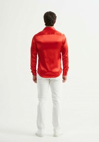 Chemise en satin rouge à manches longues avec une texture lisse et des poignets à boutons, accompagnée de pantalons blancs. La chemise présente un ourlet courbé et un design de passepoil au dos.