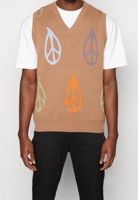 Hellbraune Strickweste mit V-Ausschnitt und mehrfarbigen Friedenszeichen in Orange, Blau, Lila und Grün. Über einem weißen Hemd getragen.