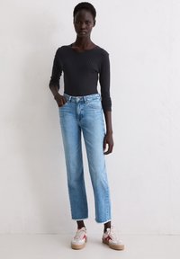 Femme se tient contre un mur blanc, portant un haut à manches longues en côtes noir, un jean droit bleu clair avec des ourlets effilochés et des baskets blanches avec des accents rouges.