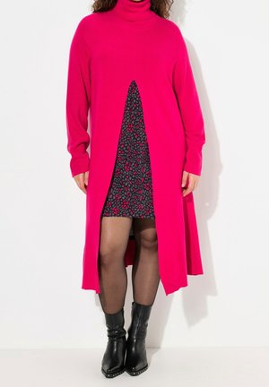 Femme portant une robe-pull à col roulé longue rose vif par-dessus une mini-robe noire et rose à motifs avec des bottines noires.