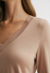 Halenka s výstřihem do V z měkkého, světlého béžového materiálu. Vyznačuje se hladkou texturou, uvolněným střihem a jemnými švy na okrajích.