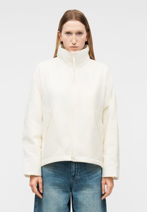 Egyenes, világosbarna hajú nő magas nyakú, fehér cipzáros dzsekit és kék farmert visel, egyszínű világos háttér előtt.