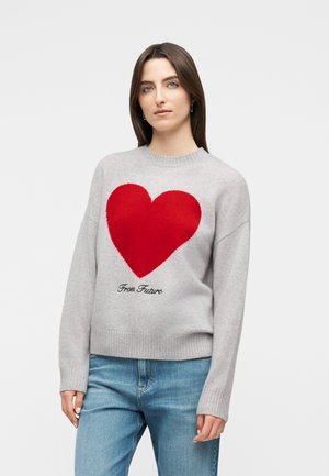 Femme portant un pull gris clair avec un grand cœur rouge et l'inscription « From Future », associée à un jean bleu, debout devant un fond uni.