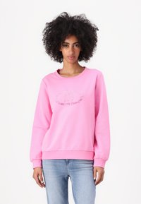 Sweatshirt rose à col rond, à manches longues et avec des poignets côtelés. Présente un logo texturé sur le devant. Porté avec un jean bleu clair.