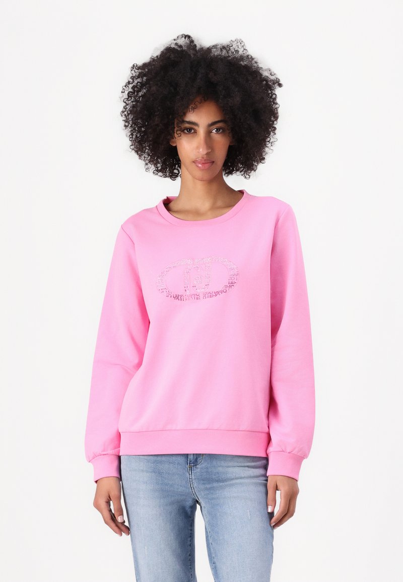 Sweatshirt rose à col rond, à manches longues et avec des poignets côtelés. Présente un logo texturé sur le devant. Porté avec un jean bleu clair.