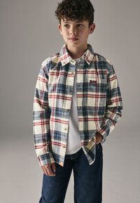 Rutet flannelskjorte i creme, marineblå og rød med knaplukning på kraven, brystlomme og gyldne knapper, båret over en hvid t-shirt.