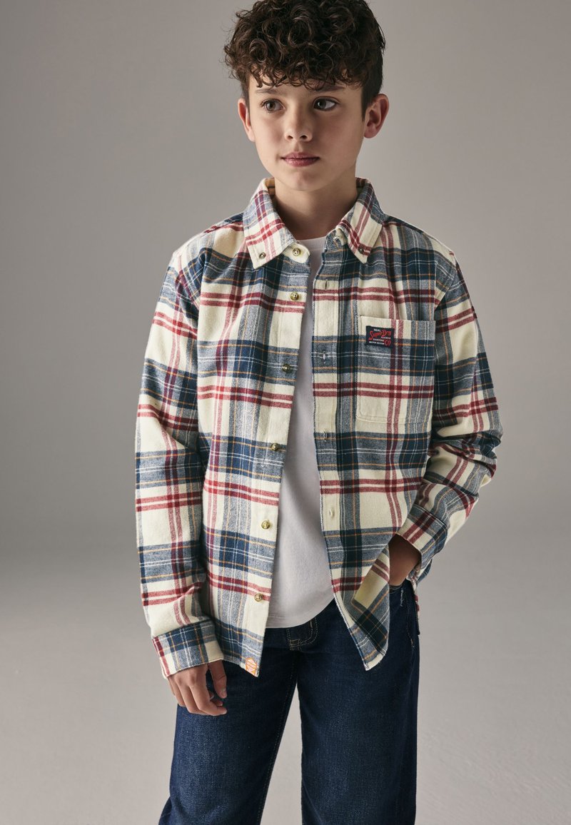 Rutet flannelskjorte i creme, marineblå og rød med knaplukning på kraven, brystlomme og gyldne knapper, båret over en hvid t-shirt.