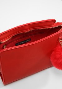 Czerwona skórzana torebka o wyraźnej, usztywnionej formie, wyposażona w zamek błyskawiczny, kieszeń wewnętrzną oraz puszysty czerwony brelok w kształcie pomponu z jednej strony.