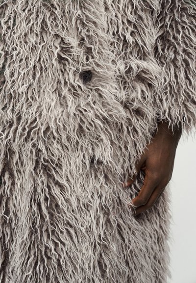 Σκούρο δέρμα χέρι που ακουμπά σε απαλό, υφασμάτινο, ανοιχτό γκρι παλτό με σγουρή γούνα και εμφανές κουμπί που ταιριάζει.