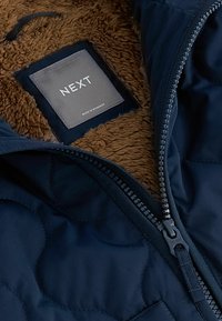 Veste matelassée marine avec doublure en polaire marron, dézippée pour montrer l'étiquette intérieure indiquant "NEXT" et "Fabriqué au Myanmar."