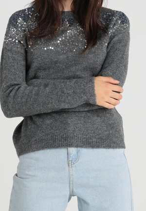 Femme portant un pull en maille gris avec des sequins argentés sur les épaules et un jean bleu clair, les bras croisés sur la poitrine.