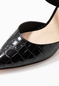 Černá lakovaná bota s špičatou špičkou, s krokodýlím vzorem a hladkým vnitřním podšitím. Otevřená po stranách, elegantní design.