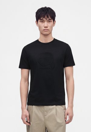Ung mand stående frontalt iført en sort t-shirt med grafik og beige bukser mod en ensartet lys baggrund.