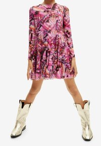 Różowa sukienka z długim rękawem, w wzór motyli, z plisowanym dołem i okrągłym dekoltem. Połączona z błyszczącymi, srebrnymi kowbojkami.