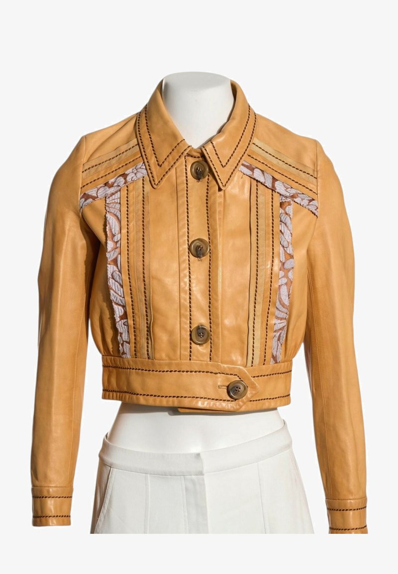 Veste en cuir courte couleur fauve avec fermeture boutonnée, détails surpiqués et panneaux en tissu fleuri sur la poitrine et les épaules.