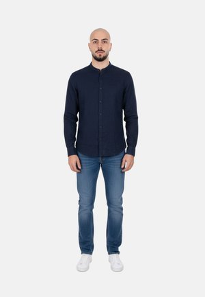 Uomo con testa rasata che indossa una camicia blu scuro con bottoni, jeans blu e scarpe da ginnastica bianche, in piedi davanti a uno sfondo bianco semplice.