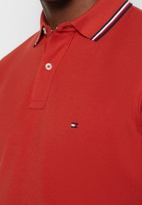 Gros plan d'un polo rouge avec un col à rayures blanches, noires et rouges, deux boutons et un petit logo de drapeau brodé sur la poitrine.