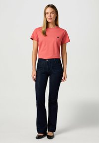 T-Shirt in Koralle mit kurzen Ärmeln und kleinem marineblauen Logo, kombiniert mit dunkelblauen Schlagjeans mit Fronttaschen und Knopfverschluss.