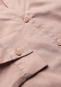 Chhemise en coton rose clair avec un détail de bouton, un tissu texturé, et un poignet soigneusement replié, avec de subtiles rayures verticales.