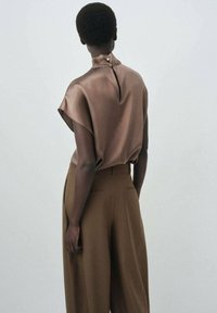 Satijnen bruine top met een hoge kraag, korte mouwen en een open rug die sluit met een knoop. Gecombineerd met losse, bruine wijde broek.