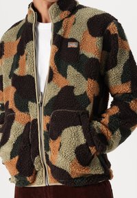 En person som bär en dragkedjefodrad fleecejacka med kamouflage-mönster, med helt äkta fickor och en liten Dickies-logotyp på bröstet, med händerna i fickorna.