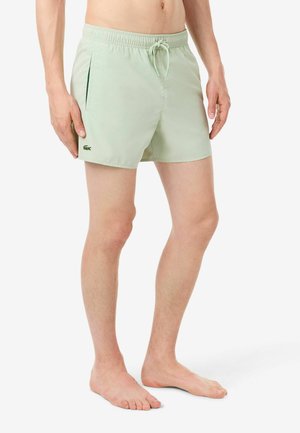 Homme portant un short de bain vert clair avec une ceinture élastique et des poches latérales, debout pieds nus sur un fond blanc.