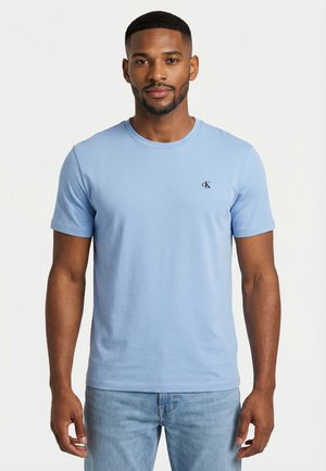 Uomo in piedi di fronte, che indossa una t-shirt Calvin Klein azzurro chiaro e jeans azzurro chiaro su uno sfondo bianco uniforme.