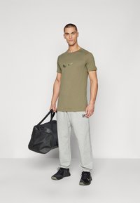 T-shirt de mangas curtas verde-oliva com um logo preto, calças de treino cinza claro e sapatilhas de atletismo pretas, segurando uma mochila de ginásio preta.