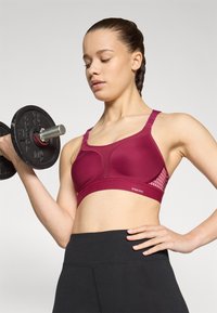 Burgunder sportsbh med mesh-detaljer, justerbare stropper og en glatt tekstur, kombinert med svarte høytlivs leggings. Modell som løfter en vekt.