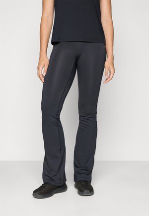Woman wearing black flared athletic pants, black sneakers, and a black sleeveless top, standing against a plain light background.
