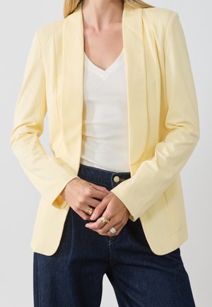 Žena v bledě žlutém saku přes bílý top s výstřihem do V a tmavě modré džíny, ukazuje ruce s několika prsteny.