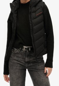Person med en svart vadderad väst över en svart tröja, med högmidjade gråtvättade jeans och ett svart bälte med en silverfärgad spänne.