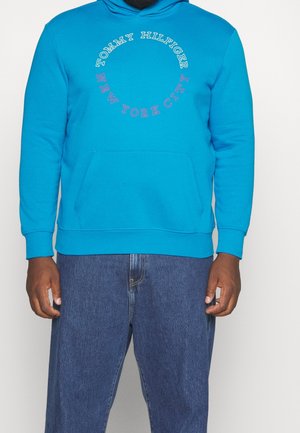 Man draagt een felblauwe Tommy Hilfiger hoodie met de tekst „New York City“ in een rond ontwerp en blauwe spijkerbroek.